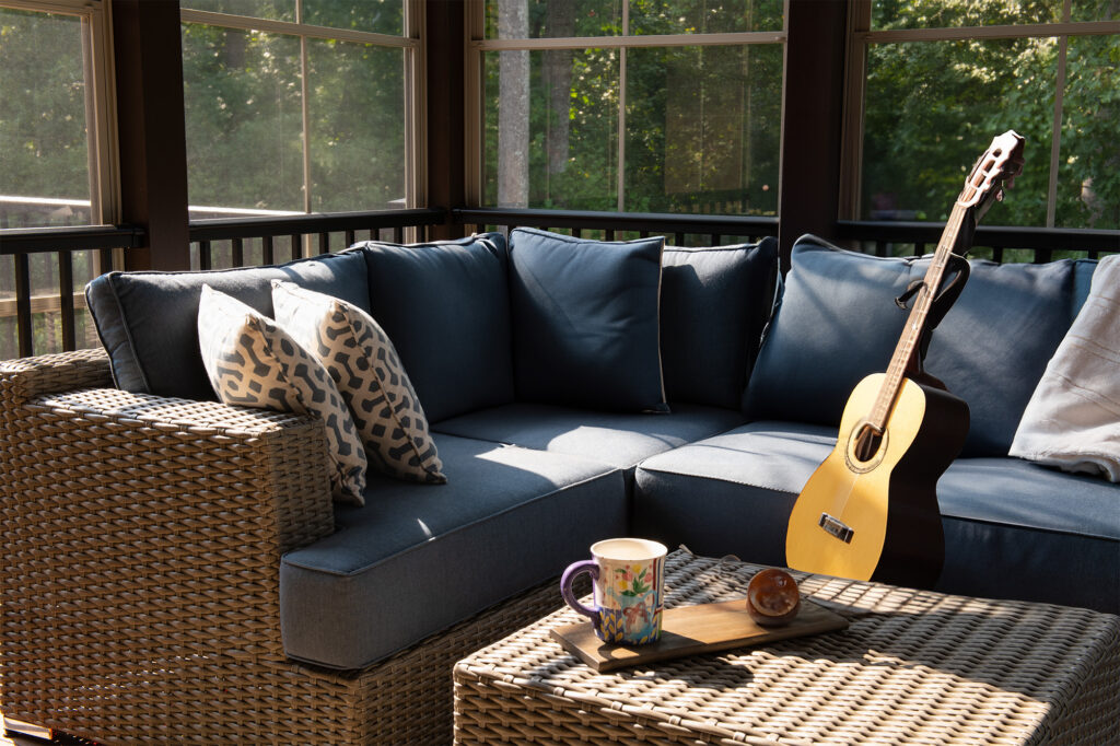 A cozy corner of an outdoor living space with modern porch windows, patio furniture, cushions and pillows. Coffee or tea pot and cups on coffee table, woods in the background.