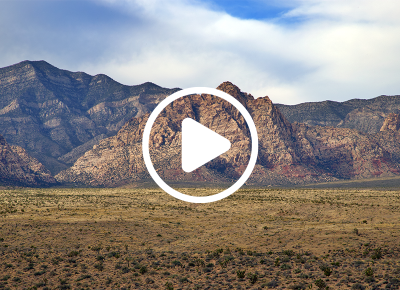 Video play button overlayed on Nevada landscape background, there is beautiful mountains with the dessert landscape and baby blue sky