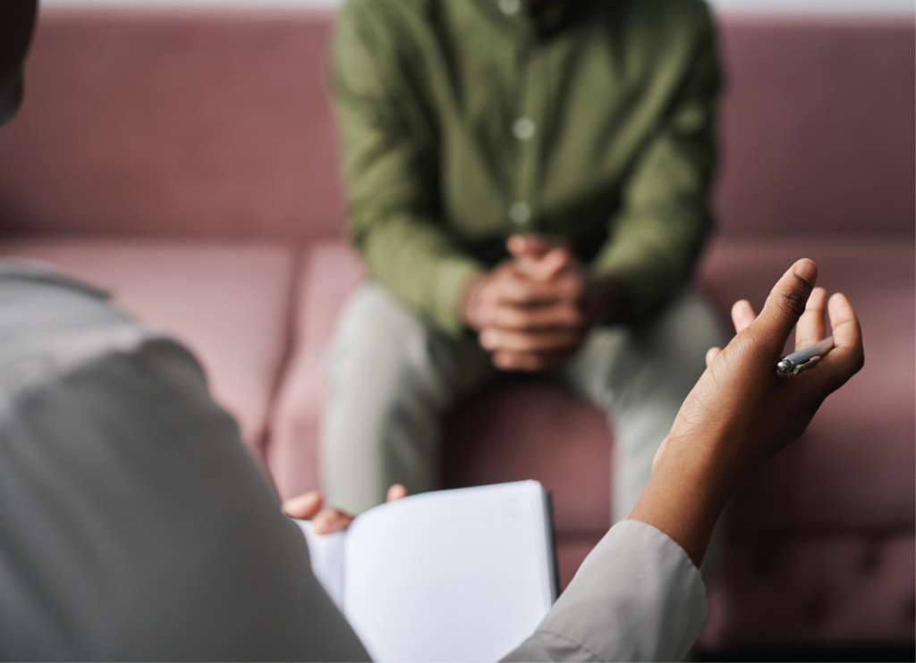 Two people talking in a therapist setting. The professional holds a pen and note book and client clasps their hands together whilst sitting forward on a washed-out maroon couch.