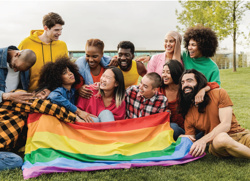 Group of queer and BIPOC folks sit together at a park with LGBTQIA+ flag and they embrace in joyous laughter and smiles.