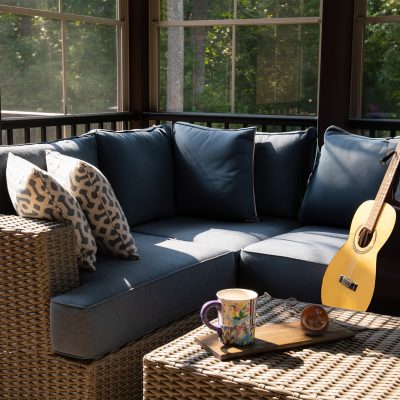 A cozy corner of an outdoor living space with modern porch windows, patio furniture, cushions and pillows. Coffee or tea pot and cups on coffee table, woods in the background.