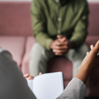 Two people talking in a therapist setting. The professional holds a pen and note book and client clasps their hands together whilst sitting forward on a washed-out maroon couch.