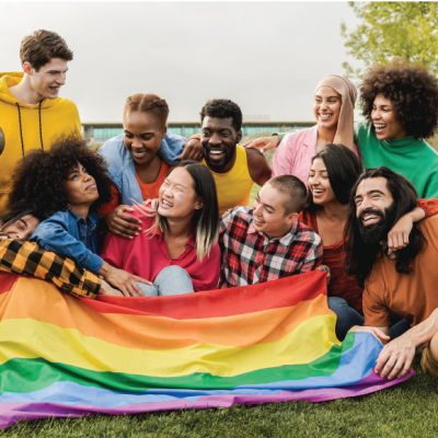 Group of queer and BIPOC folks sit together at a park with LGBTQIA+ flag and they embrace in joyous laughter and smiles.